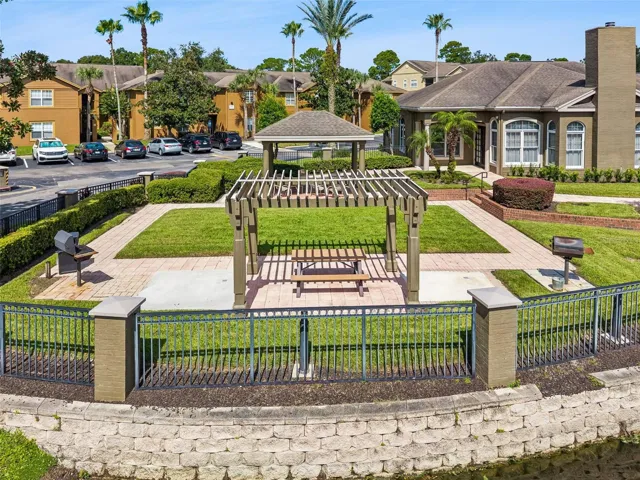 Sabal Point pergola with a picnic table and barbecue grills on a brick paver patio beside the lake wall, with the clubhouse in the background.