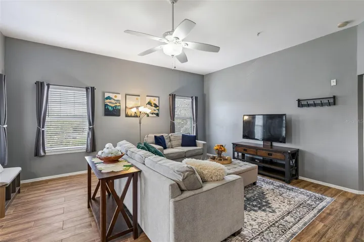 Living room with two gray sofas, an ottoman, TV console, ceiling fan, and large windows with blinds and curtains.