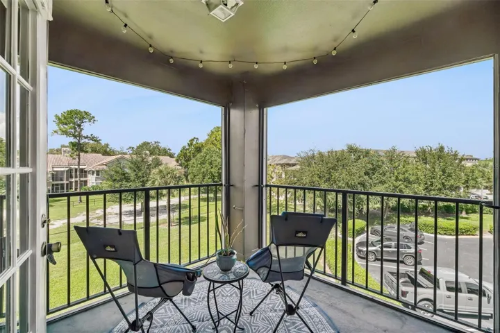 Screened balcony with two chairs, a small table, and string lights, overlooking green space and a parking area below.