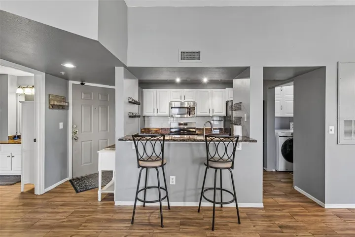View from the entry into the open kitchen with granite breakfast bar, white cabinets, stainless microwave, and the laundry closet visible at right.