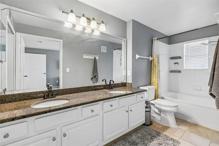 Primary bathroom with a double-sink granite vanity, large wall mirror, vanity lighting, toilet, tub and shower combination, and tile flooring.