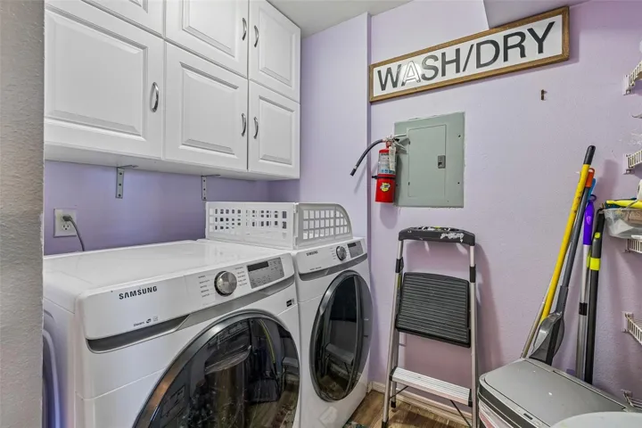 Laundry closet with a Samsung front-load washer and dryer, upper white cabinets, shelving, and the electrical panel.