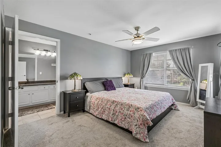 Primary bedroom with a king bed, carpeted floor, ceiling fan, window with curtains, and a doorway to the en suite bathroom.