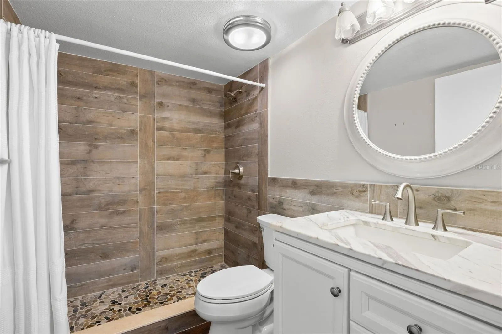 The updated second bath features a white vanity, oval mirror, and a tiled walk-in shower with wood-look walls and a pebble-style floor.