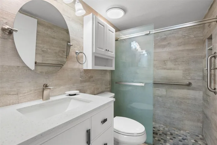 Updated in 2021, the primary ensuite features a sleek white vanity, large round mirror, and a walk-in shower with wood-look tile, pebble-style flooring, frameless glass, and grab bars.