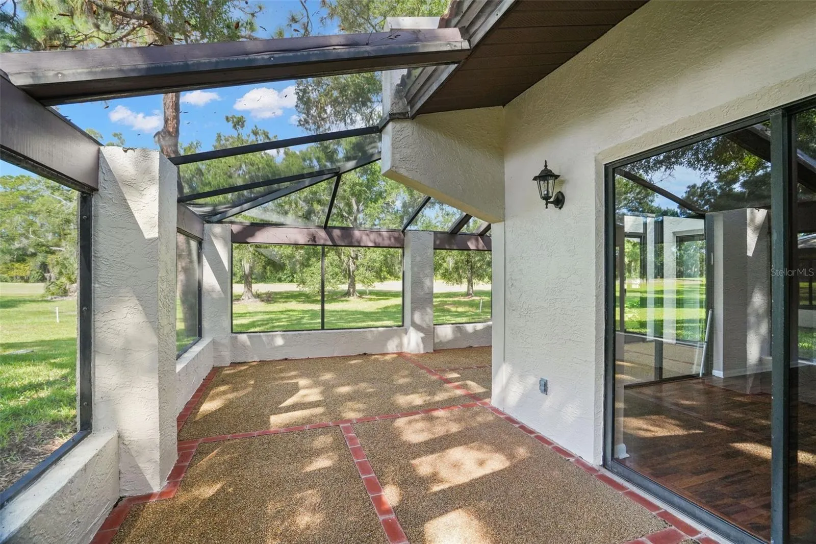 The oversized screened lanai extends the living space outdoors, with room for separate seating and dining areas beneath a combination of covered and screened sections. Brick accents edge the textured flooring, while mature trees and open green space create a peaceful backdrop.