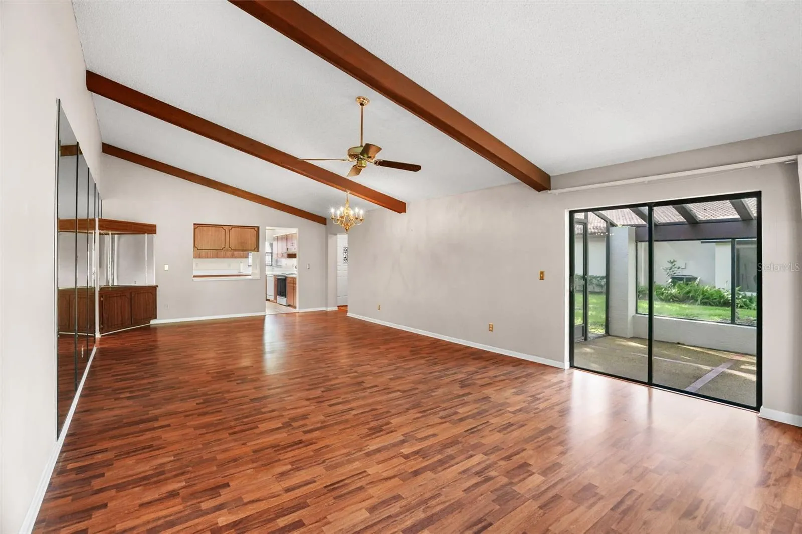 An open living and dining area offers plenty of room to arrange furniture your way, with vaulted ceilings, exposed wood beams, warm-toned laminate flooring, and sliding glass doors opening to the screened lanai. A pass-through to the kitchen keeps the spaces connected without giving up definition.
