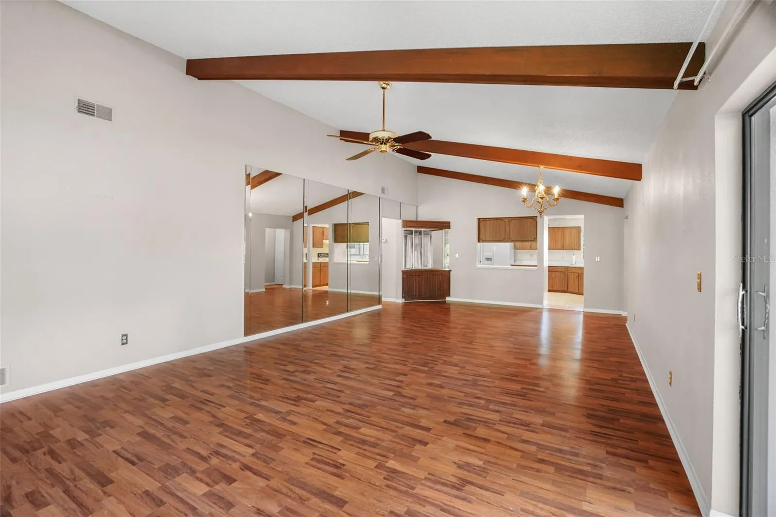 The long, open layout gives you plenty of flexibility for both everyday living and dining, with vaulted ceilings, exposed wood beams, a ceiling fan, and warm laminate flooring. A mirrored wall reflects light across the room, while built-in cabinetry near the dining area adds convenient storage and serving space.