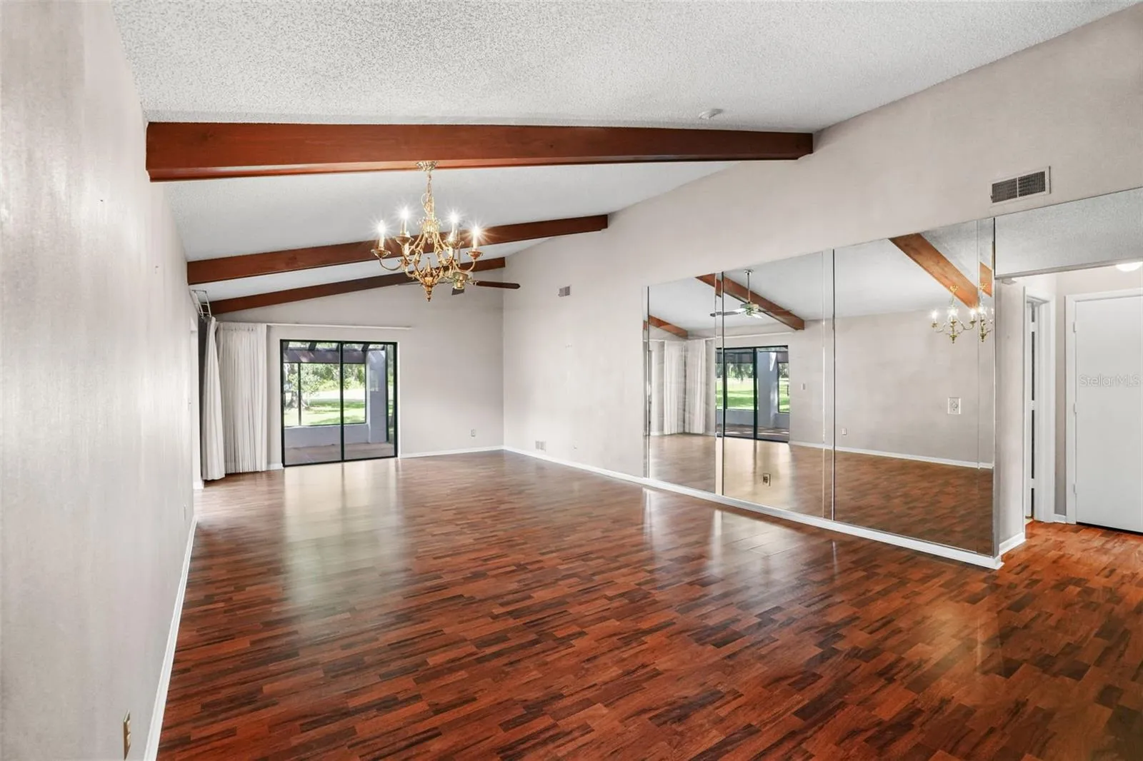A vaulted ceiling with exposed wood beams brings warmth and architectural interest to the spacious living and dining area. Rich laminate flooring runs throughout, while a full mirrored wall reflects natural light and sliding glass doors open directly to the screened lanai.