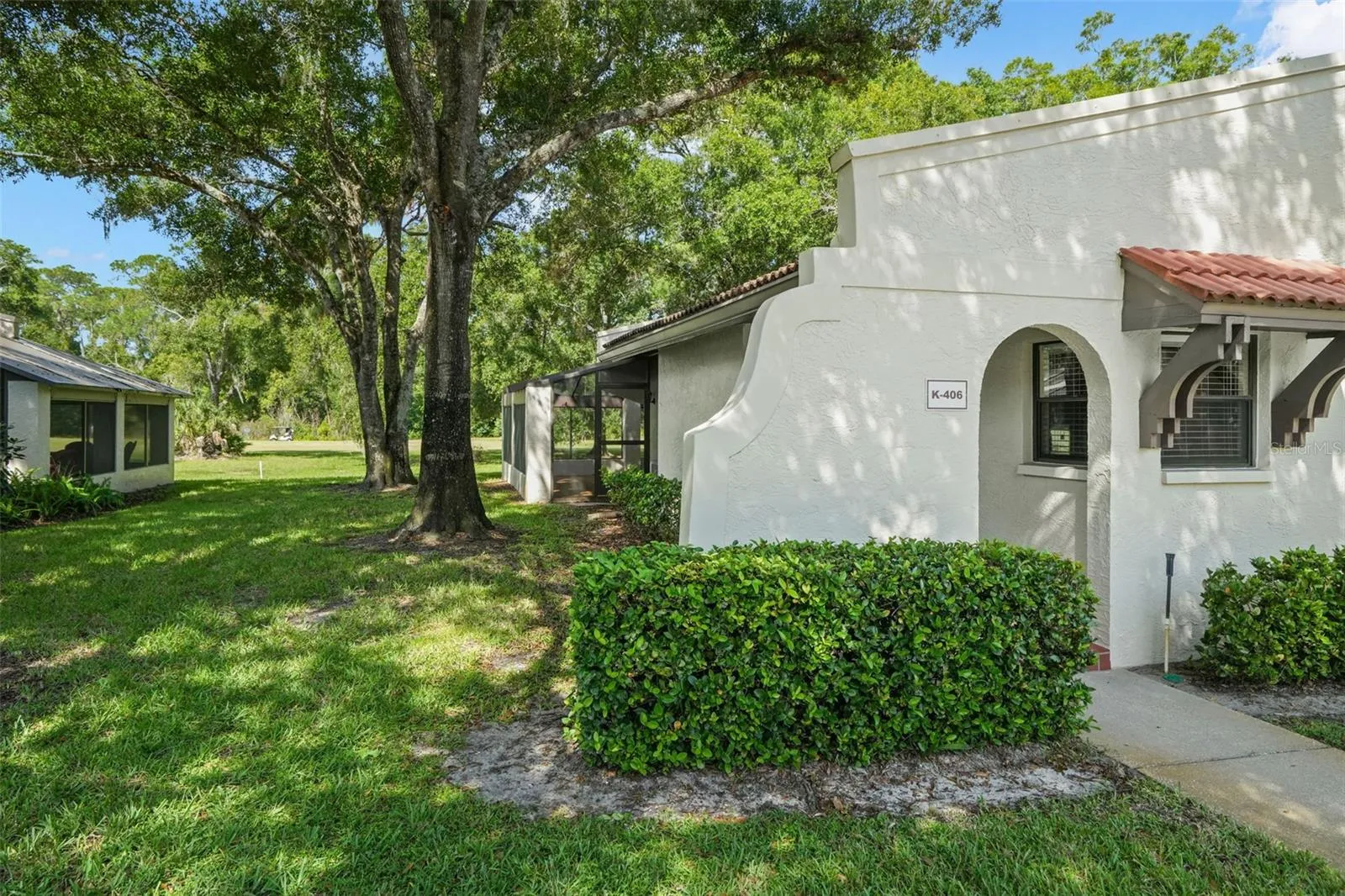 The corner location opens up the setting with mature shade trees, lawn along the side, and a clear view toward the screened lanai. The private entry is tucked beneath the home’s arched stucco detailing, while the extra side exposure brings more natural light into the condo.