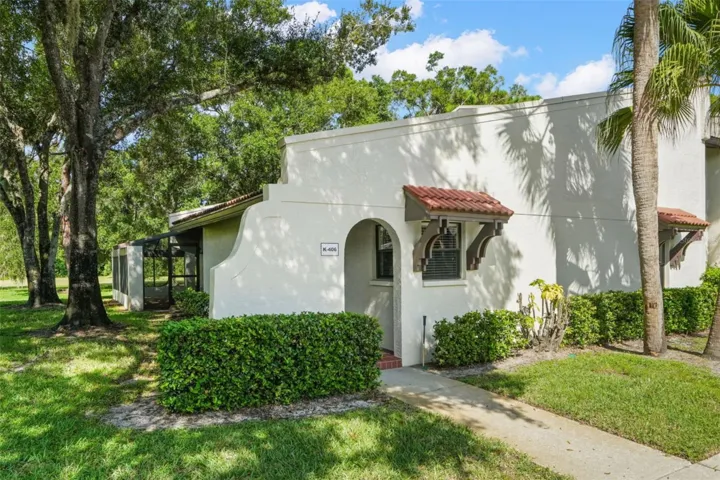 A first-floor corner location gives this 2 bedroom, 2 bath condo a private entrance, no stairs to navigate, and extra green space along the side. Mature oaks and palms frame the stucco exterior, while red tile accents add a touch of Mediterranean character; the screened wraparound lanai can be seen extending toward the rear.