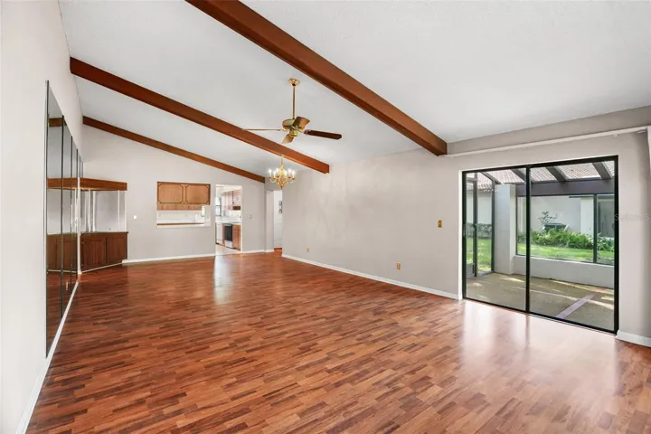 An open living and dining area offers plenty of room to arrange furniture your way, with vaulted ceilings, exposed wood beams, warm-toned laminate flooring, and sliding glass doors opening to the screened lanai. A pass-through to the kitchen keeps the spaces connected without giving up definition.