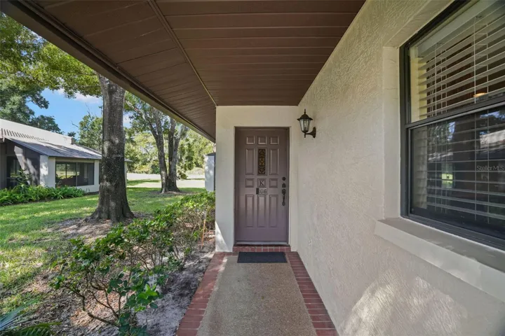 A covered walkway leads to the front door, edged in brick and shaded by mature trees. A large front window brings light into the condo, while the end-unit location keeps the entry feeling more open and tucked away from passing foot traffic.