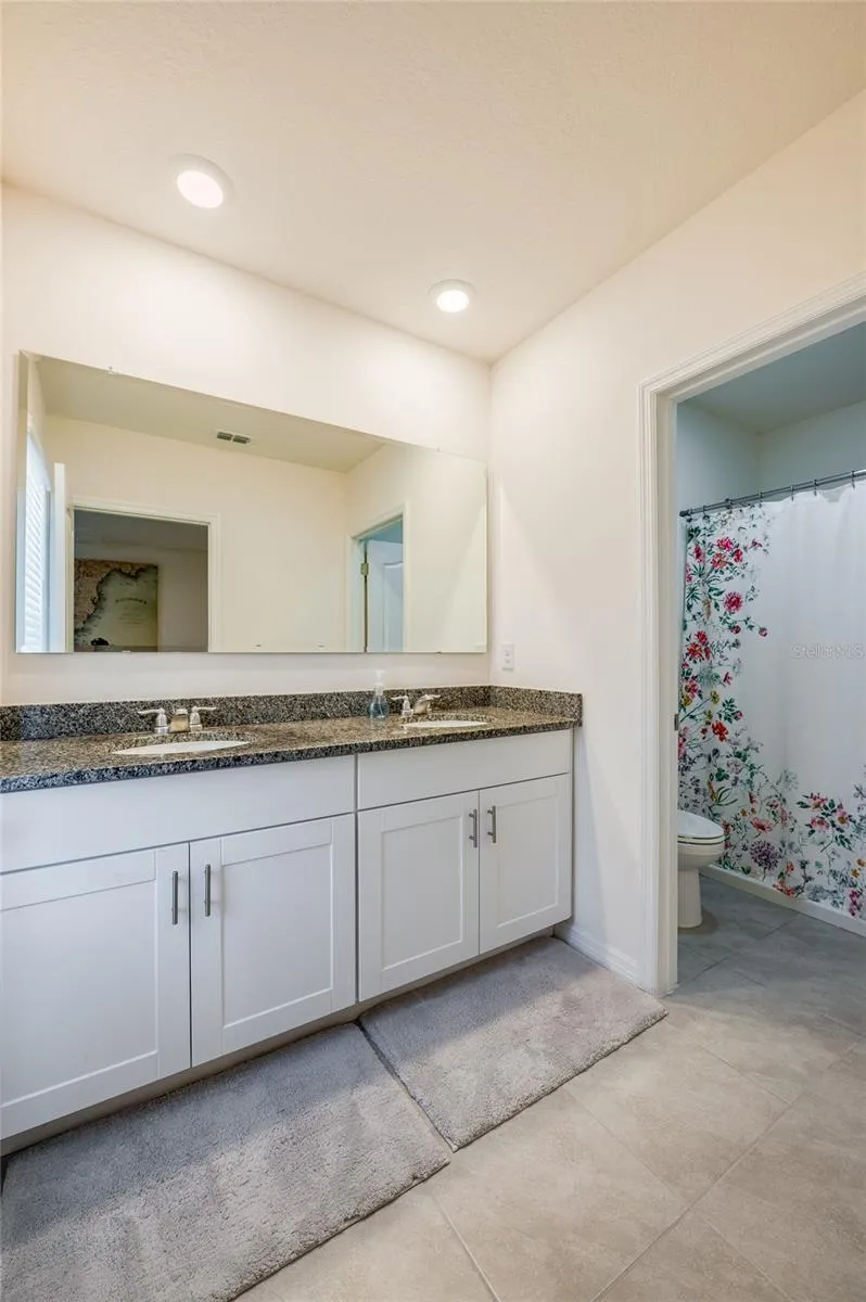 Bright bathroom vanity area features dual sinks, granite countertop, white cabinetry, a large wall mirror, and recessed lighting.