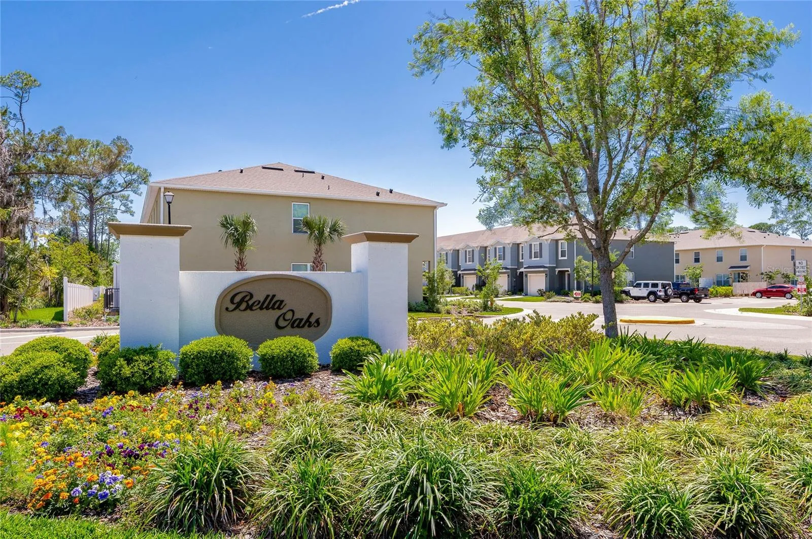 Landscaped Bella Oaks community entrance features mature greenery and colorful plantings.