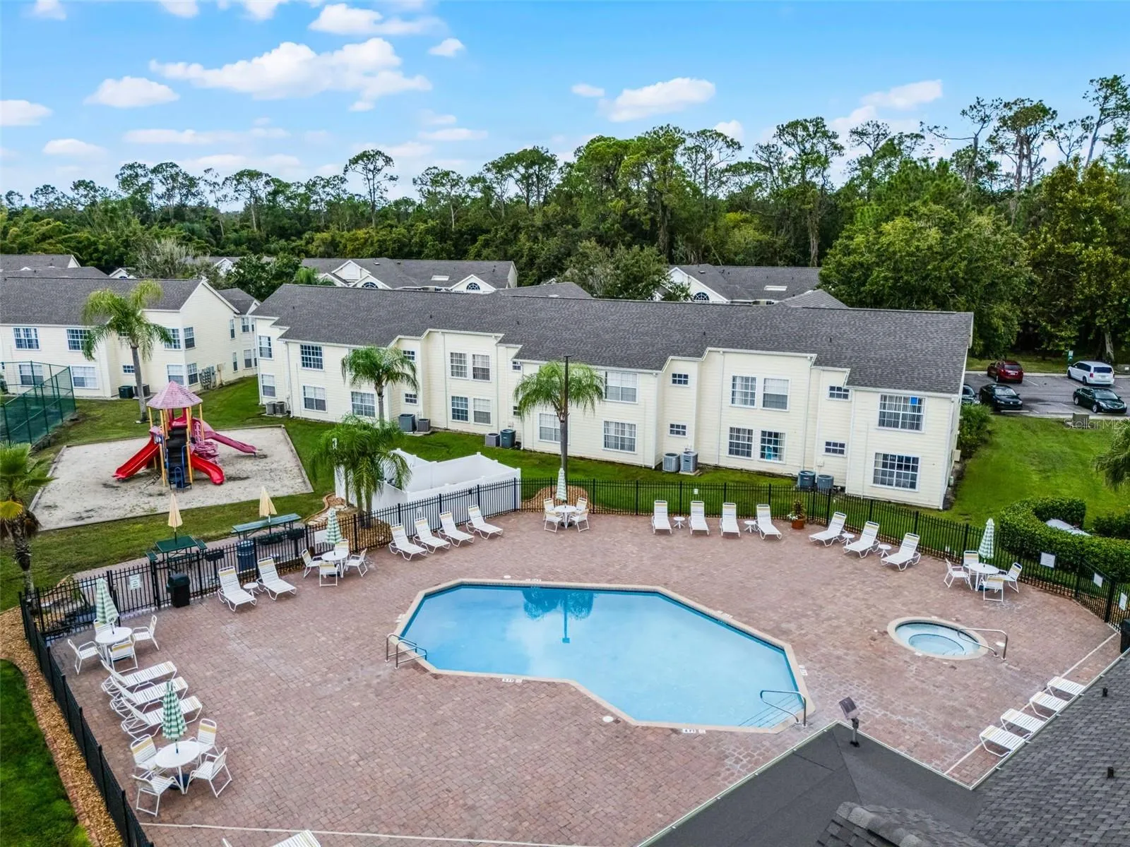 Resort Pool &amp; Playground View: Angled drone view of the paver pool deck, integrated hot tub, lounge seating, fenced perimeter, and adjacent children's playground.