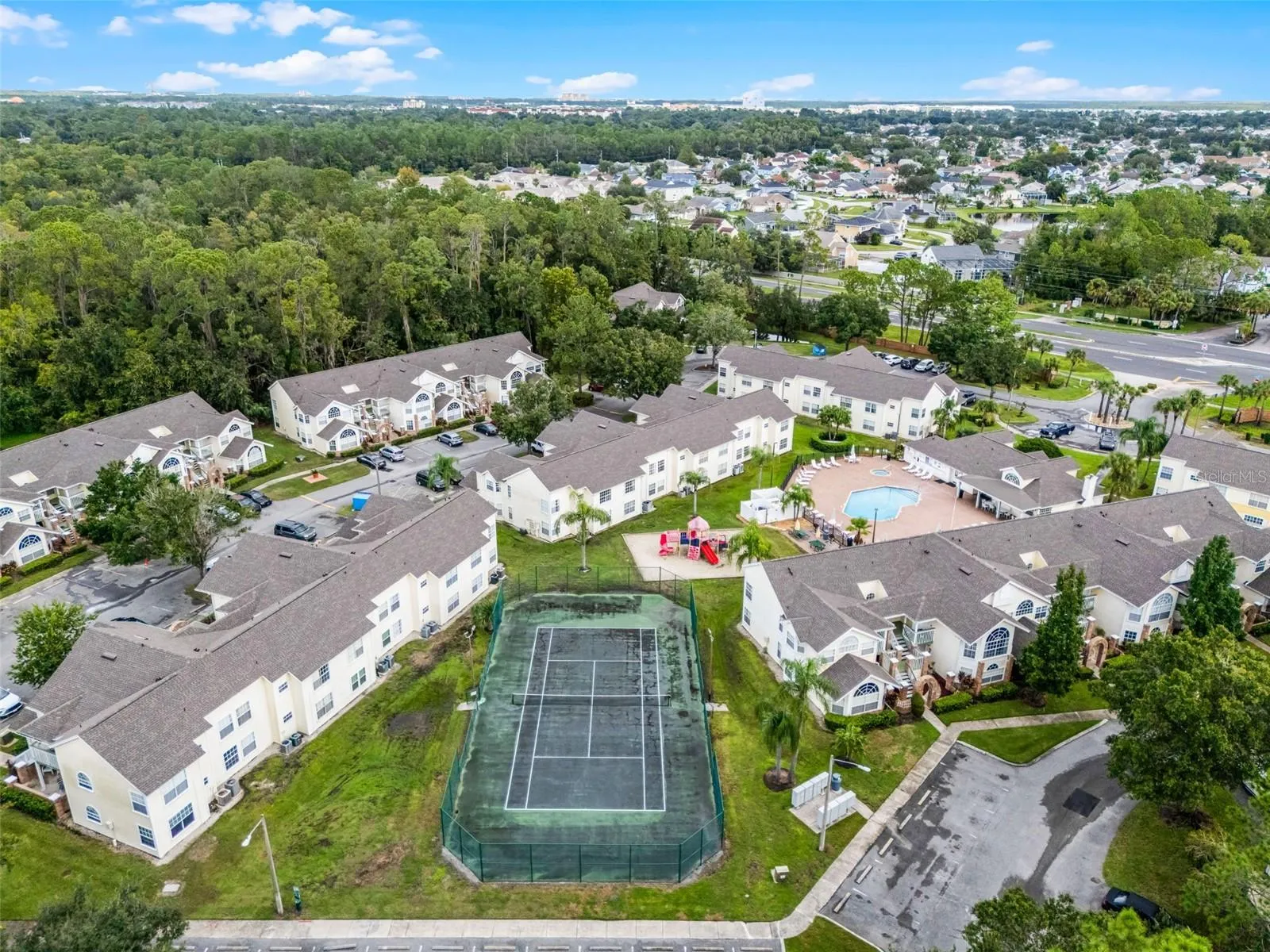 Community Amenities Aerial: High-angle drone shot displaying key community features, including the fenced tennis court, children's playground, resort pool, clubhouse, and surrounding residential buildings.