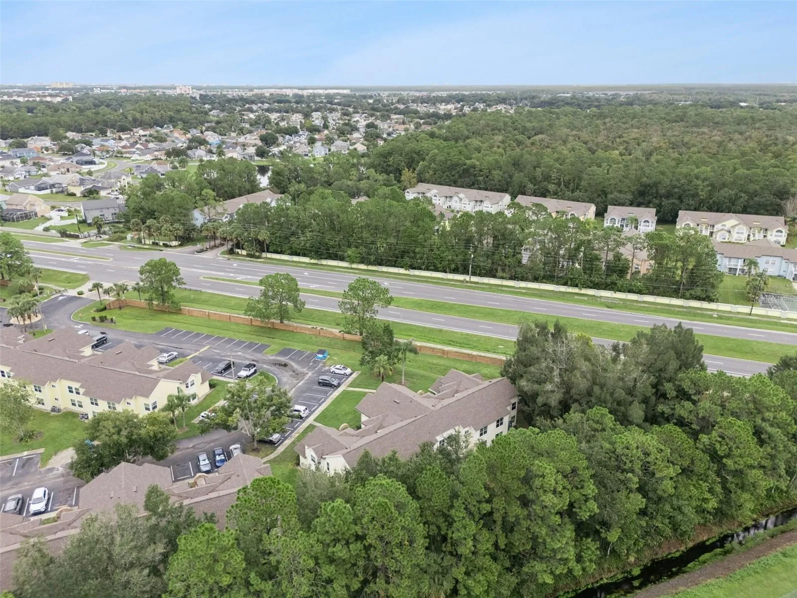 High-Angle Regional Aerial: Elevated drone view displaying the building nestled beside a lush pine tree buffer, showing parking access, surrounding neighborhoods, and convenient proximity to major roadways.