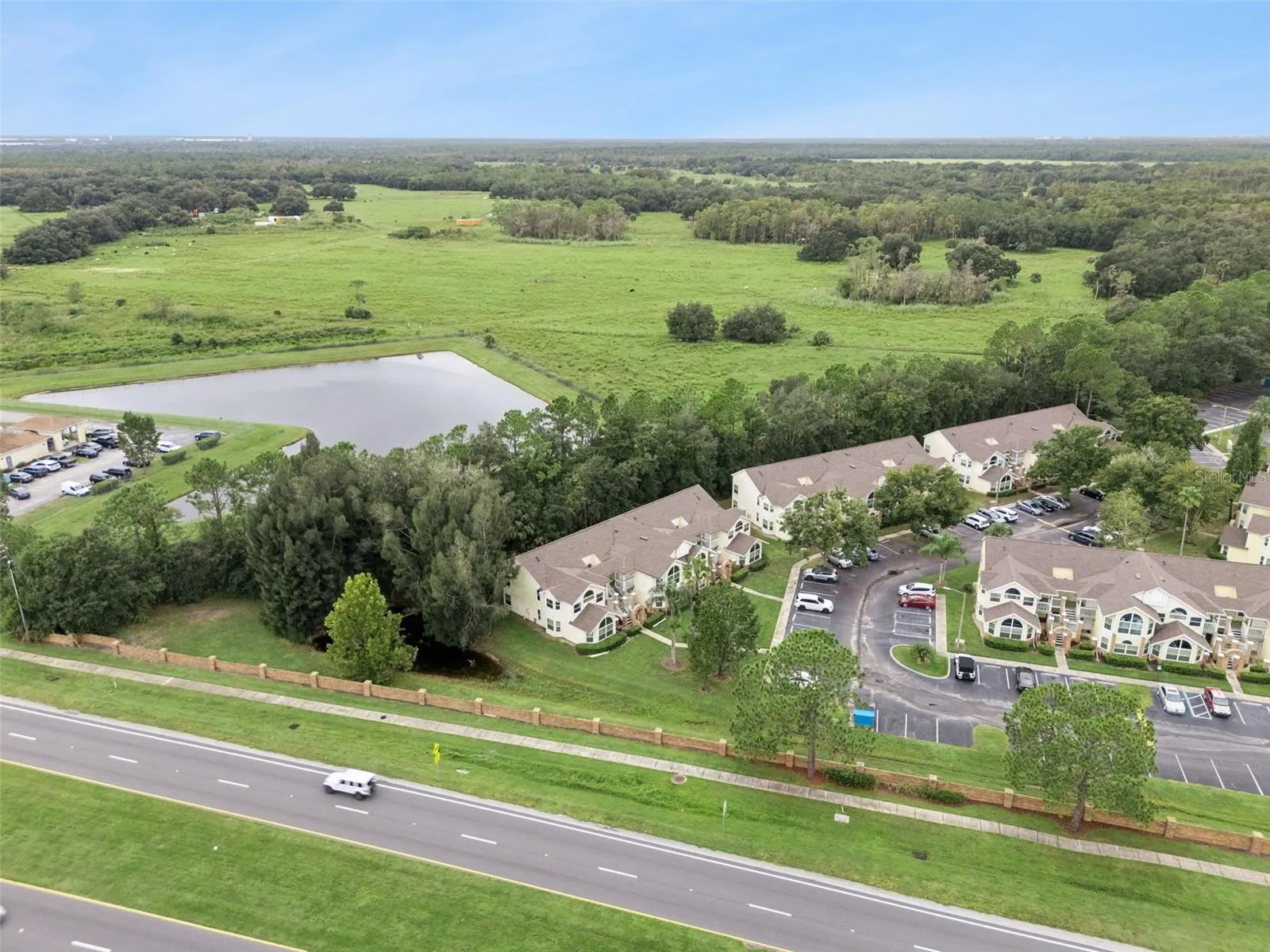 Water Feature &amp; Open Fields Aerial: Wide aerial perspective featuring the community's adjacent tranquil pond, wooded buffer line, and rolling green backdrop.