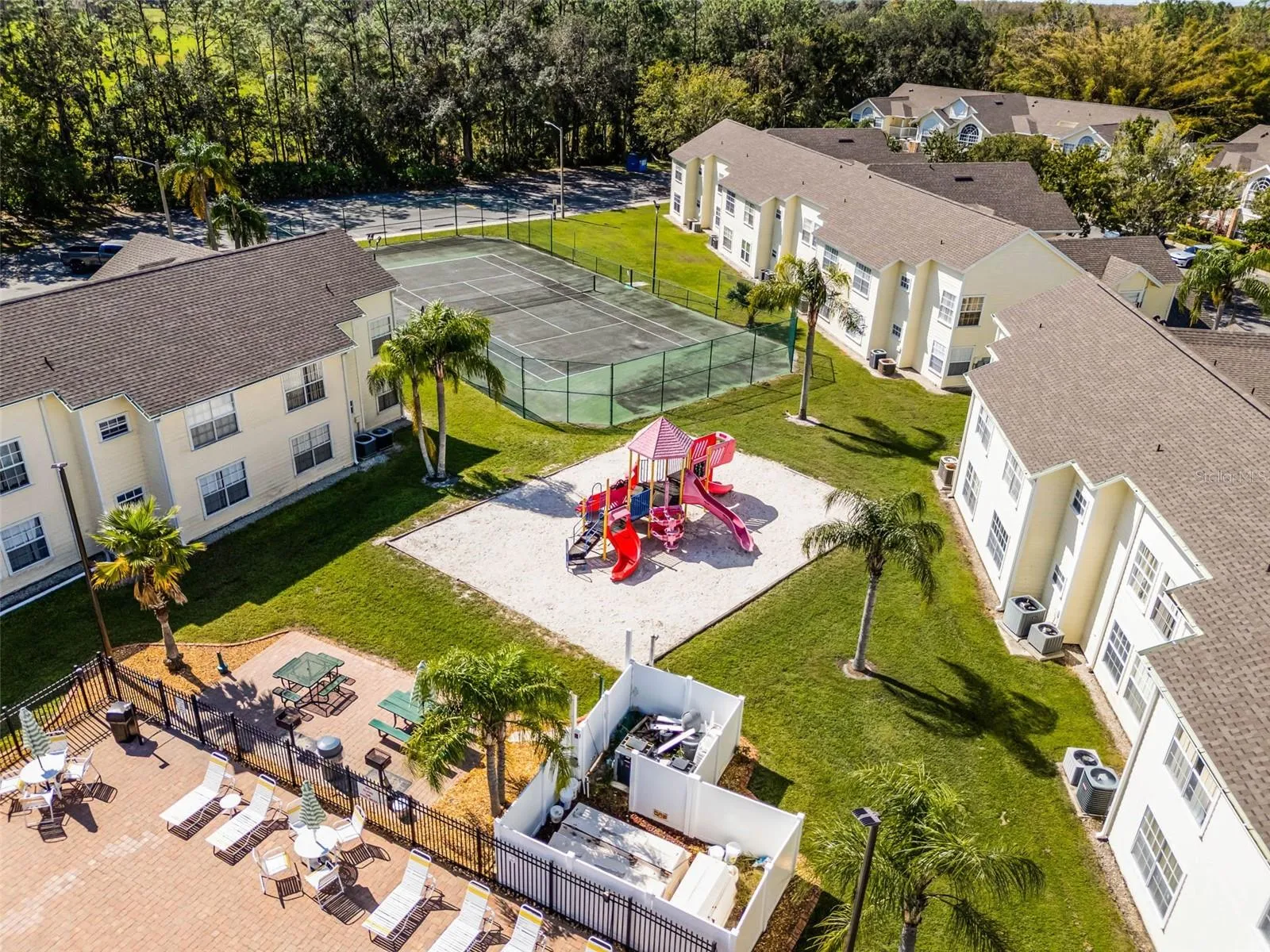 Outdoor Recreational Areas: Focused aerial view showcasing the community playground structure, picnic/grilling zone, fenced tennis court, and manicured green grounds.