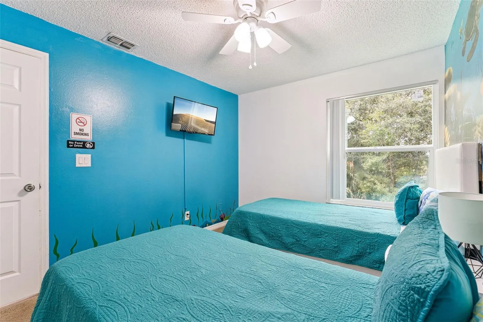 Ocean-Themed Guest Bedroom: Bright guest bedroom featuring vibrant ocean blue accent walls, dual twin beds, a ceiling fan, and wall-mounted TV.