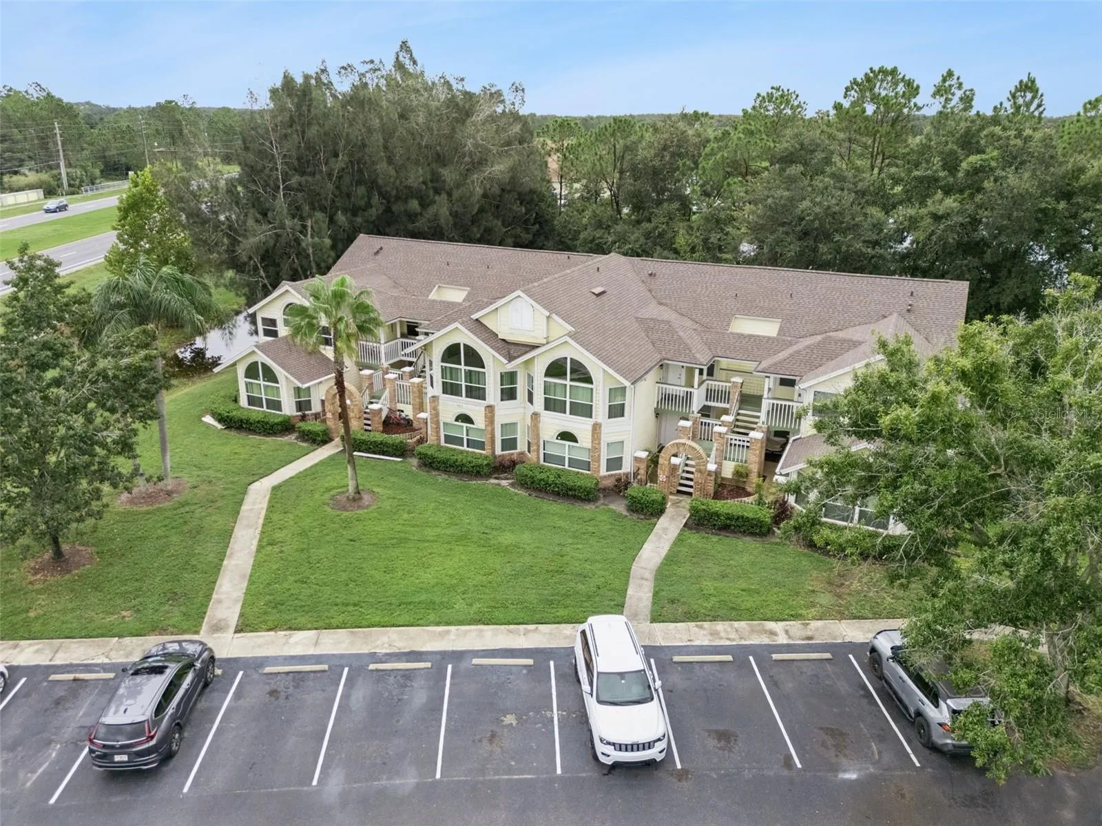 Building Facade Aerial: Focused front aerial capture highlighting the exterior facade, brick-accented entryways, central lawn, and parking access.