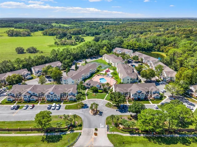 Community Entrance &amp; Layout Aerial: Broad aerial view capturing the main entrance roundabout, central pool deck, clubhouse, tennis court, and residential layout.