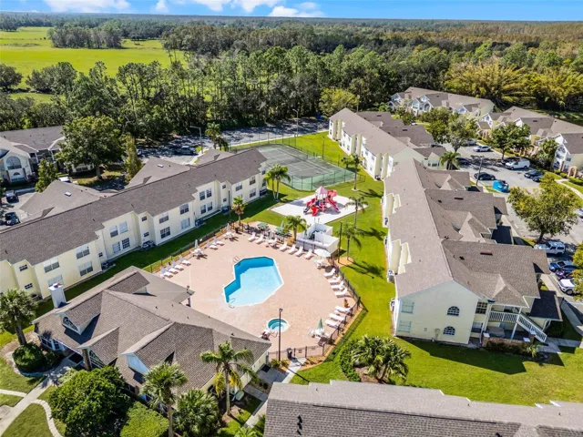 Amenity Hub Aerial: Elevated aerial view highlighting the central pool deck, clubhouse, sand playground, tennis court, and surrounding residential buildings set against open green spaces.
