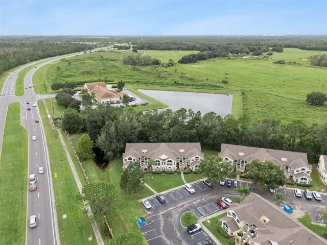 Community &amp; Pond Overview: Broad aerial panorama displaying the condominium grounds, nearby water feature, surrounding open green spaces, and adjacent thoroughfare.