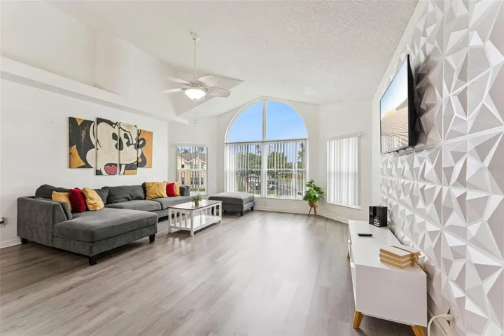 Living Room: Sun-drenched living room boasting vaulted ceilings, a geometric 3D feature wall, wood-look plank flooring, and a large architectural arched window