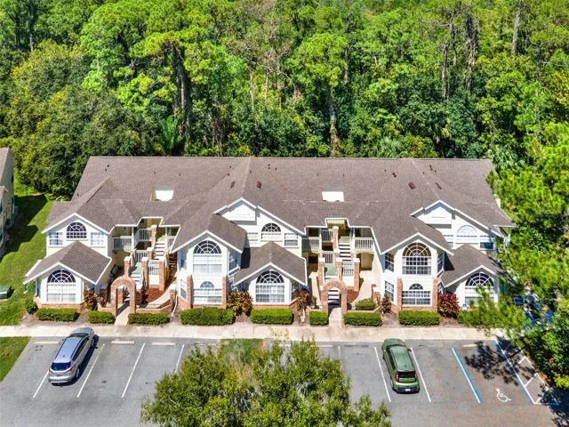 uilding Exterior Aerial: Direct front aerial perspective showcasing the unit building, parking spaces, brick entry archways, and dense wooded tree canopy behind.