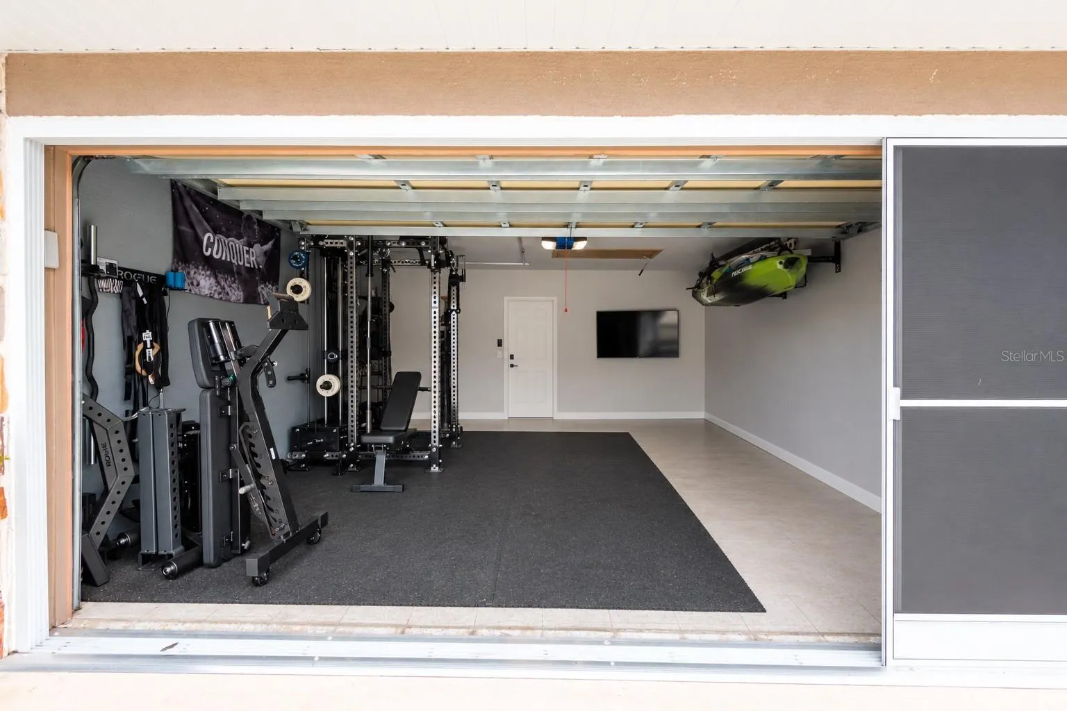 Garage with a screen and virtually removed items except for the exercise area, TV and kayak attached to the ceiling. Painted walls with tile flooring.
