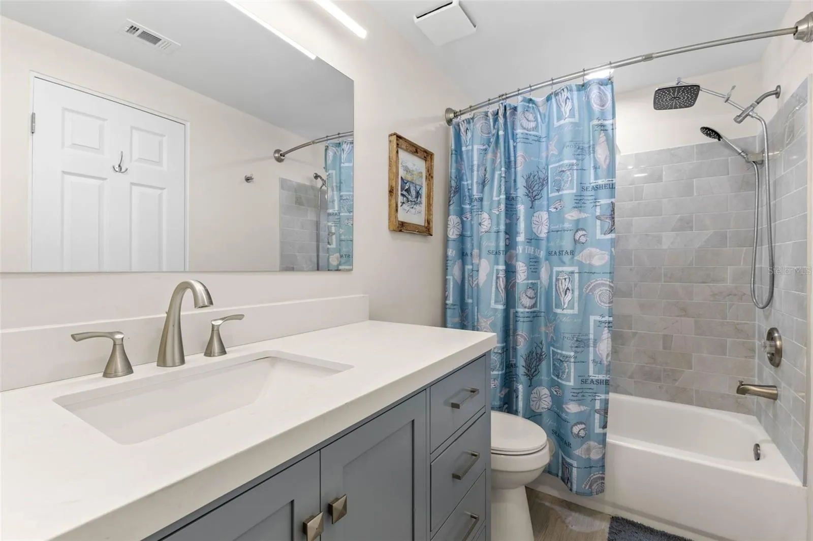 The remodeled primary bath (2025) pairs a wide gray vanity and sleek white countertop with a tiled tub/shower surround, rainfall showerhead, handheld sprayer, and updated fixtures for a clean, modern finish.