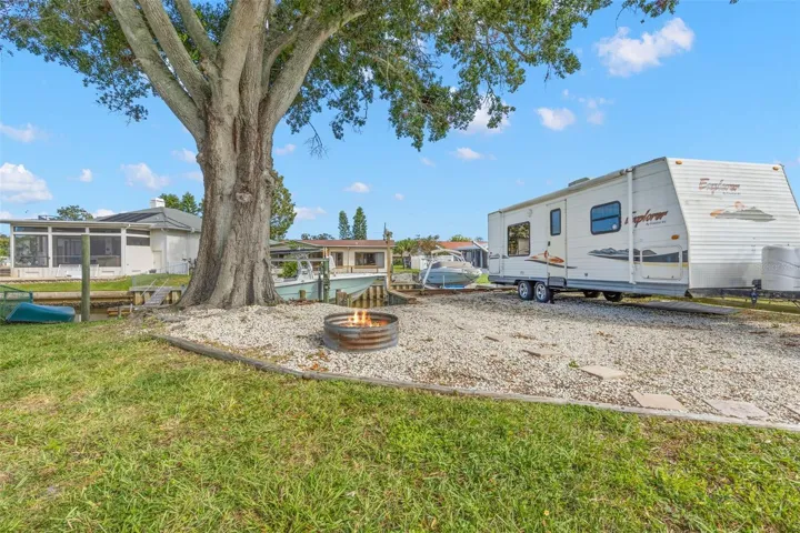 The extended side yard adds exceptional flexibility with room for RV, boat, or trailer parking, while a gravel gathering area with a fire pit sits beneath a massive shade tree just steps from the canal.