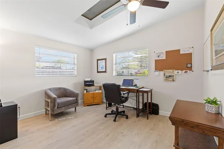 A bright secondary bedroom, currently used as a home office, offers two windows, a skylight, ceiling fan, neutral tones, and luxury vinyl plank flooring (2025), with plenty of flexibility for guests, work, or hobbies.
