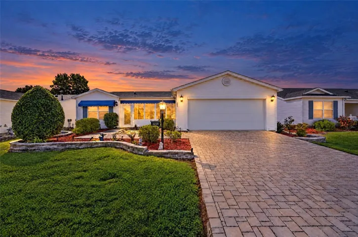 Beautiful twilight view showcasing the home's attractive landscaping, decorative stone retaining walls, expansive brick-paver driveway, blue awnings, and welcoming exterior lighting.