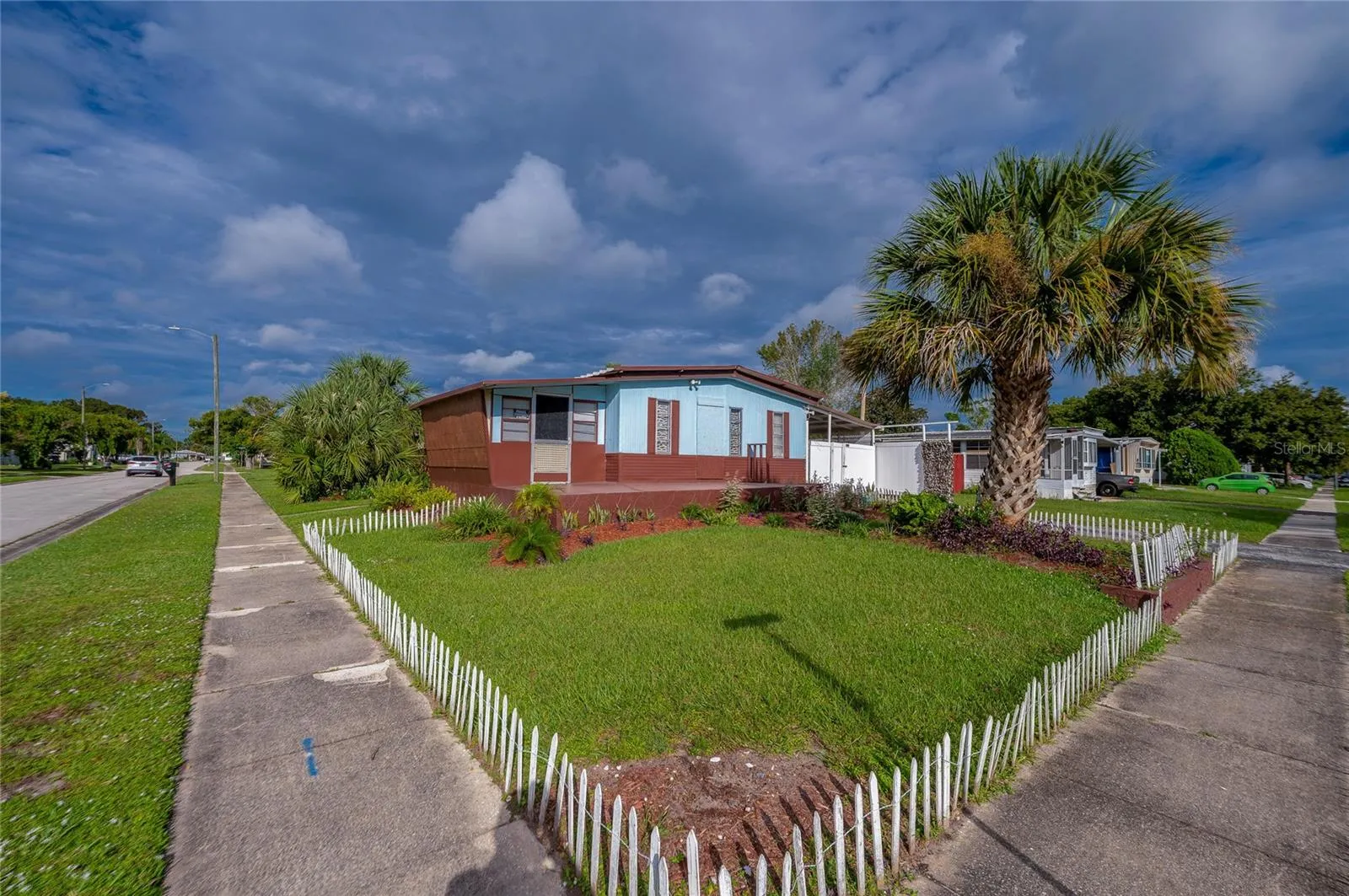 Wide exterior view highlights the corner setting, lawn, tropical landscaping, and low decorative fencing surrounding the front yard.