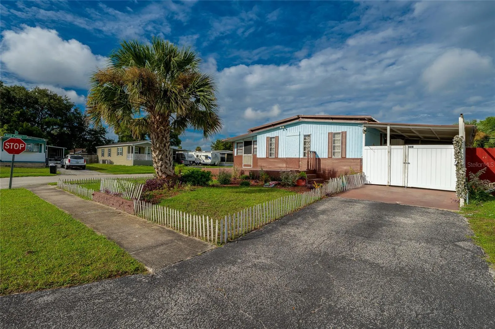 Front view showcases the home’s landscaped yard, mature palm tree, driveway, covered parking area, and welcoming entry.