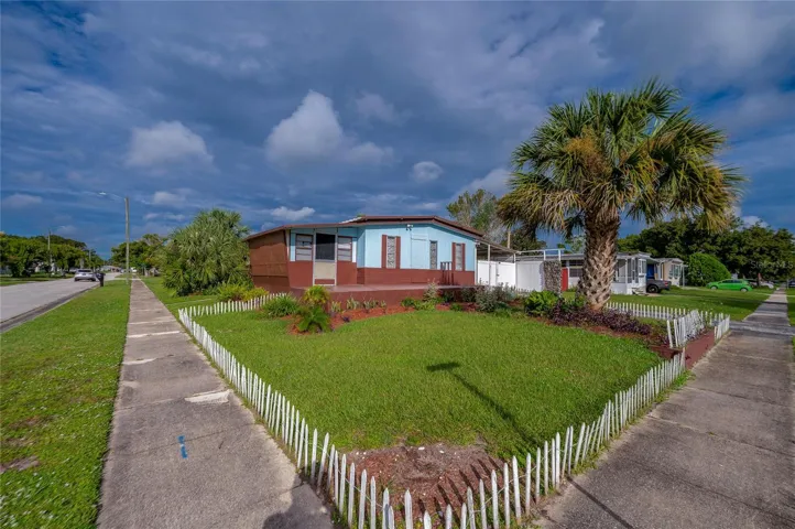 Wide exterior view highlights the corner setting, lawn, tropical landscaping, and low decorative fencing surrounding the front yard.