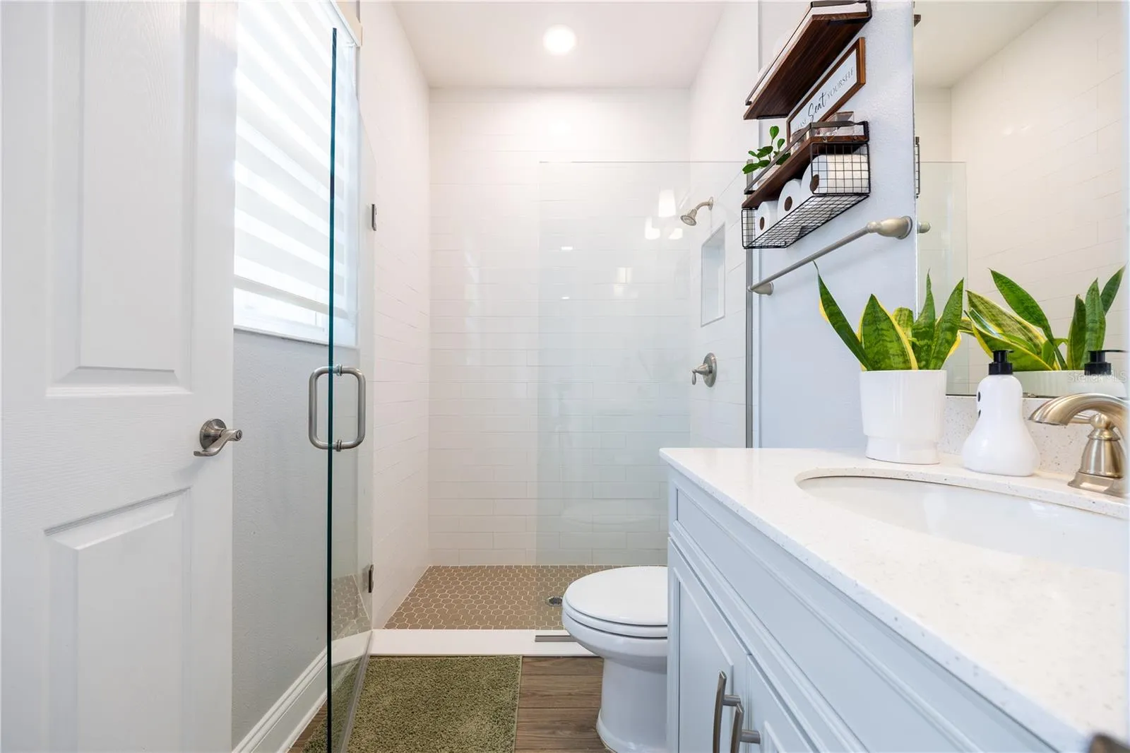 Bright bathroom featuring a frameless glass shower, white tile walls, mosaic shower floor, and a spacious vanity countertop.