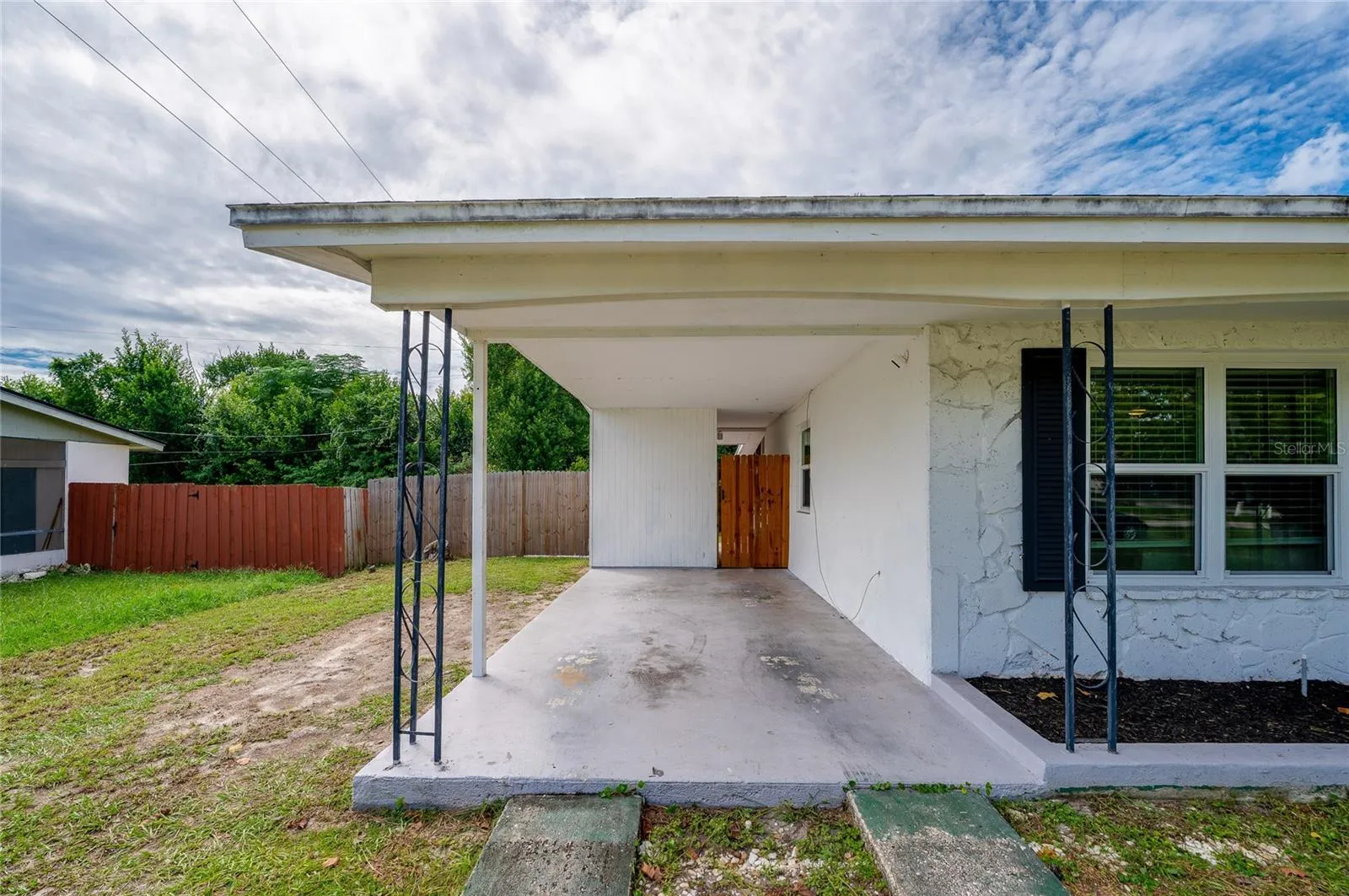 Covered concrete carport with wrought-iron support posts, a textured exterior wall finish, and a gated side pathway.