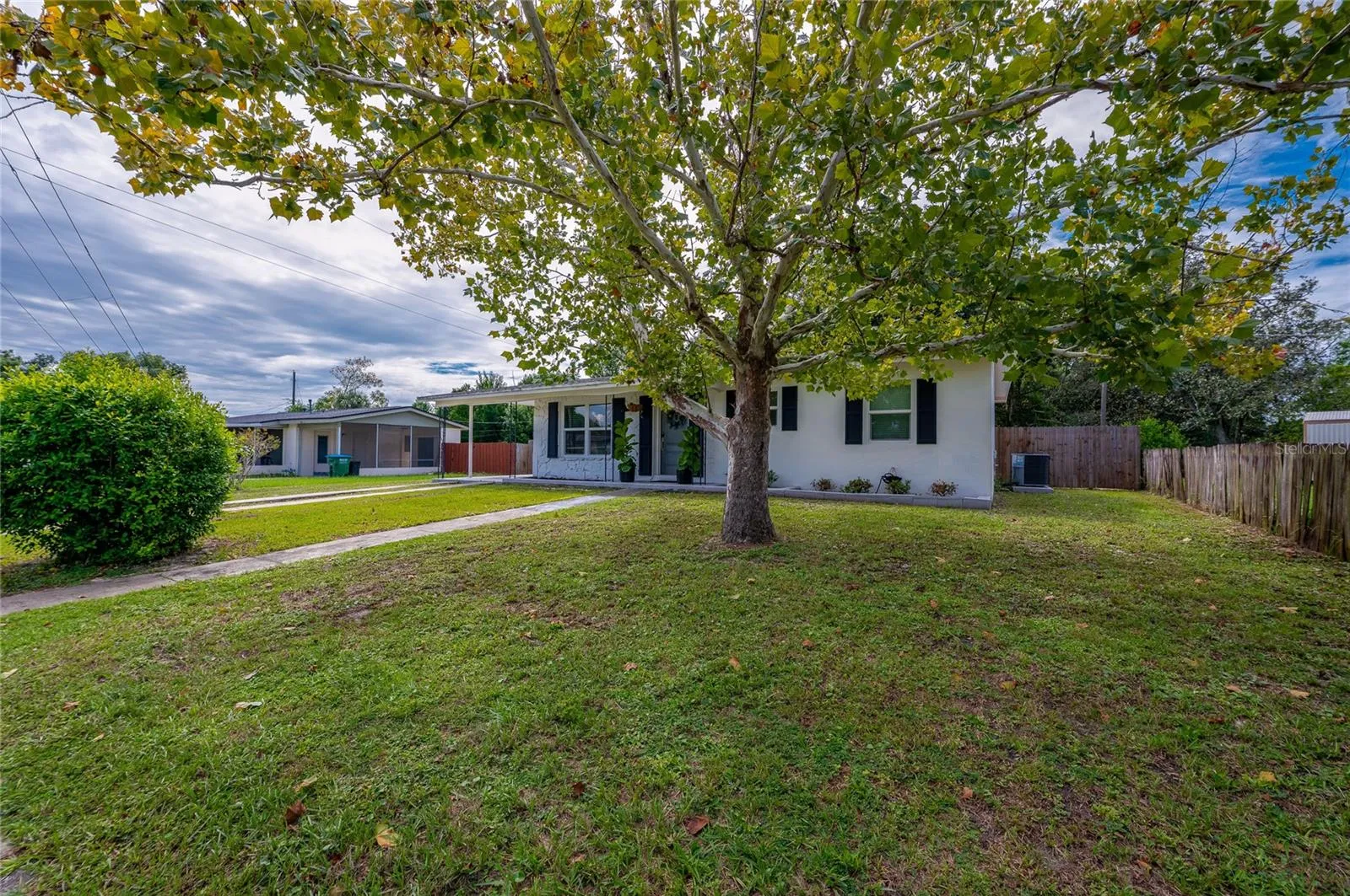Lush front yard shaded by a large leafy tree, featuring a paved walkway leading to the home's single-story facade with dark window shutters.