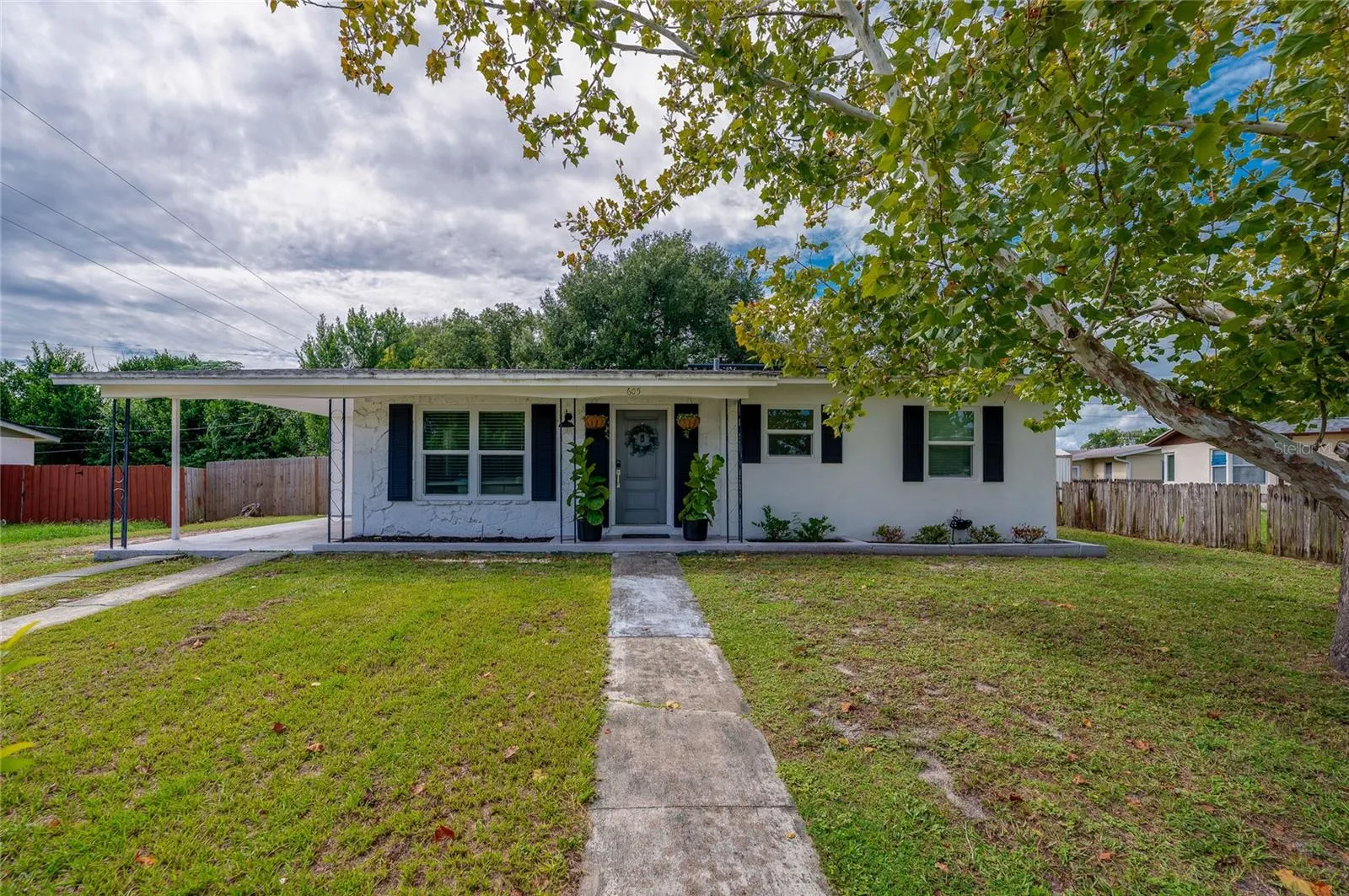 Charming single-story home exterior featuring crisp white siding, dark accent shutters, a covered carport, and a paved walkway crossing a spacious front lawn.