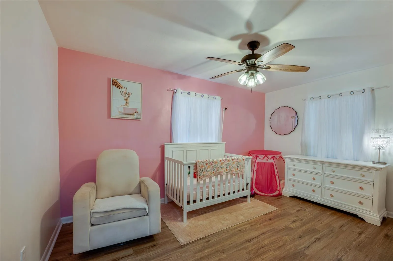 Bright bedroom area highlighted by warm LVP flooring, soft pink accent walls, a ceiling fan, and ample natural light from two windows.