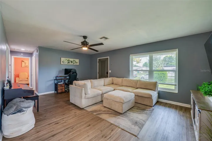 Generously sized main room featuring LVP flooring, soft gray walls, a central ceiling fan, and large front-facing windows.