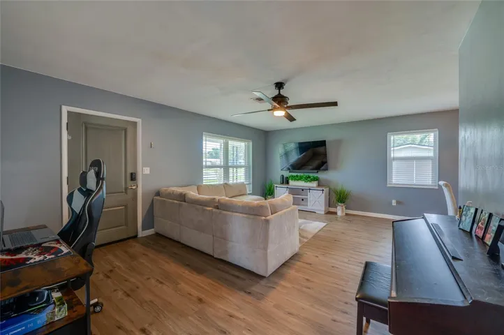 Open living area showing LVP floors, cool gray walls, a ceiling fan, and large windows that fill the space with light.