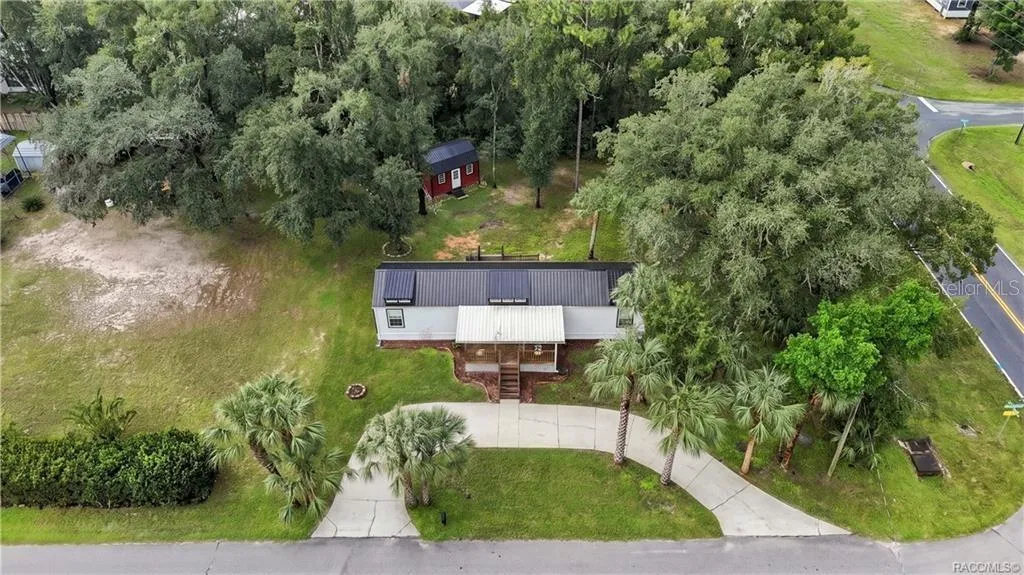 Aerial Property View – A beautiful overhead perspective of the .29-ACRE CORNER HOMESITE, showcasing the METAL ROOF, COVERED FRONT PORCH, circular concrete driveway, mature trees &amp; detached storage building.