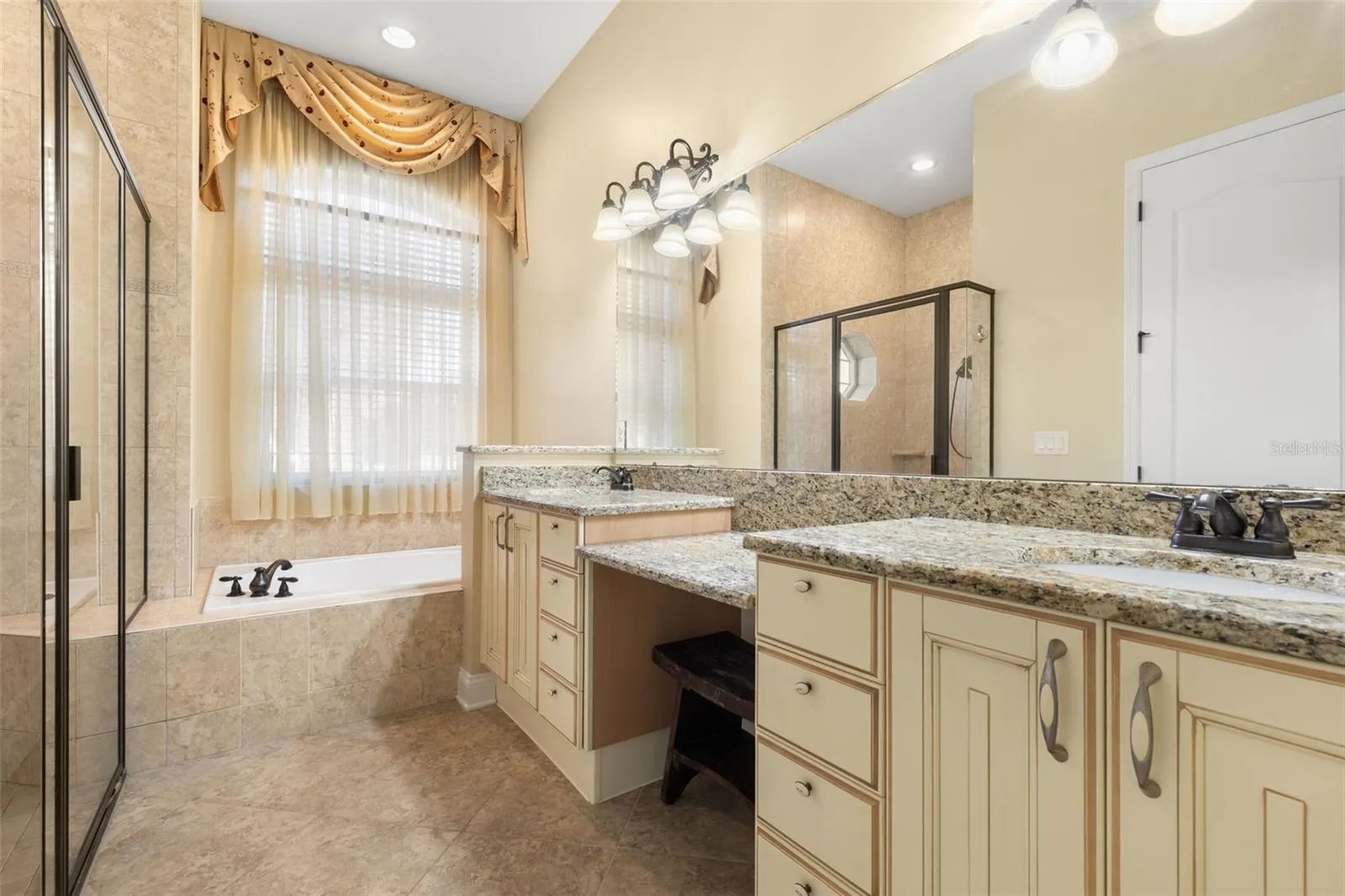 An Italian-inspired primary bathroom featuring dual vanities with granite countertops, a dedicated makeup area, soaking tub, and separate tiled shower.