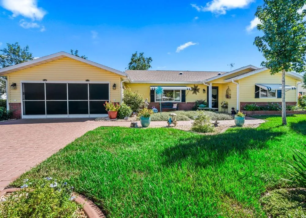 Charming front exterior with cheerful yellow siding, brick accents, a screened garage, and a brick-paver driveway. A covered front porch, striped window awnings, and colorful landscaped beds add inviting curb appeal.