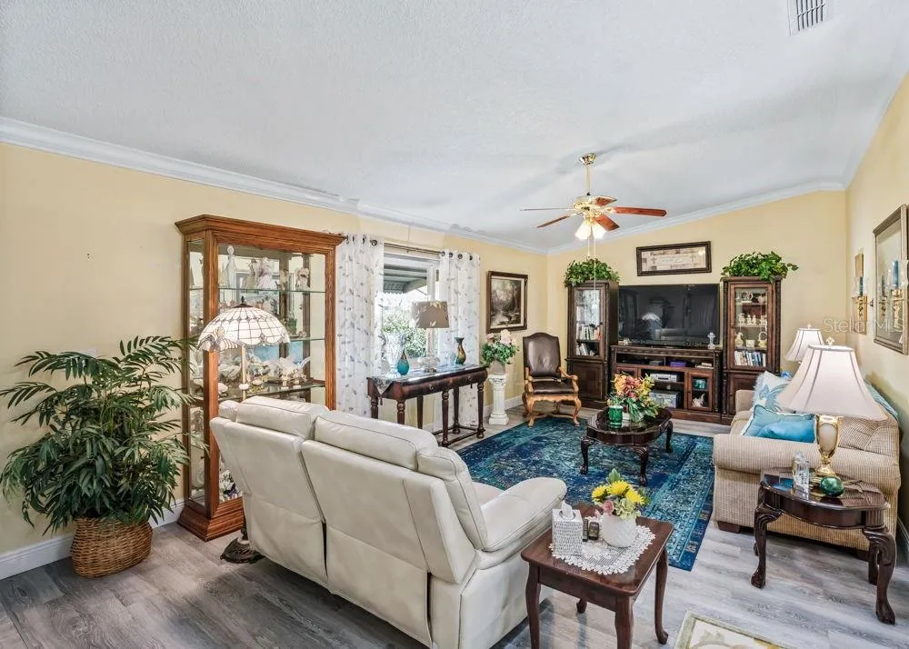 Bright and inviting living room featuring wood-look vinyl flooring, crown molding, and a ceiling fan. A large window welcomes natural light, while soft neutral walls create a comfortable setting for relaxing or entertaining.